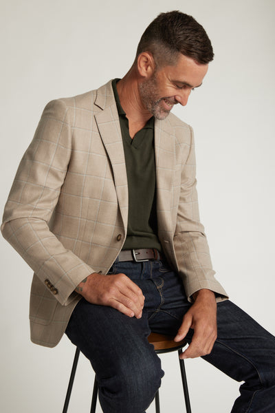 A man with short brown hair and a beard sits on a stool, smiling as he looks down. He wears the Barkers Yorkshire Check Blazer over a dark green shirt, blue jeans, and a brown belt against a plain background.