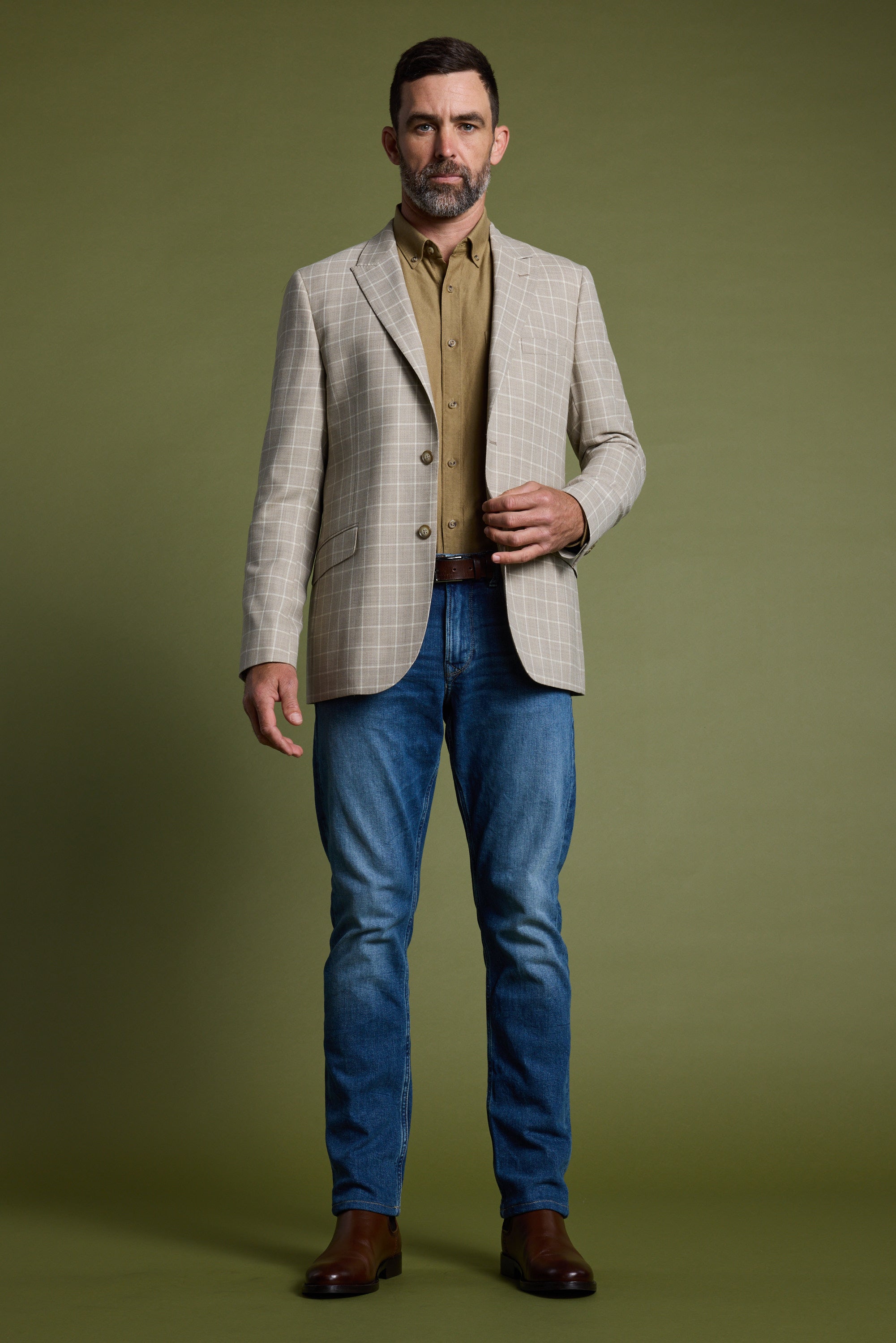A man with short brown hair and a beard stands against an olive green background, wearing the Barkers Prestwick Check Blazer, a light brown shirt, blue jeans, and brown boots.