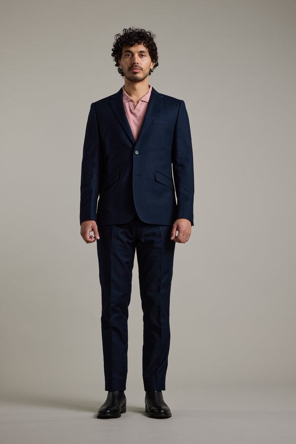 A man with curly hair stands facing forward in a Barkers Moris Cotton Blazer, a light pink dress shirt, and black dress shoes against a plain light gray background.