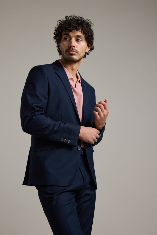 A man with curly hair models the Barkers Moris Cotton Blazer over a light pink shirt, standing against a plain background and gazing thoughtfully to his left, with one hand near his face and the other at his waist.