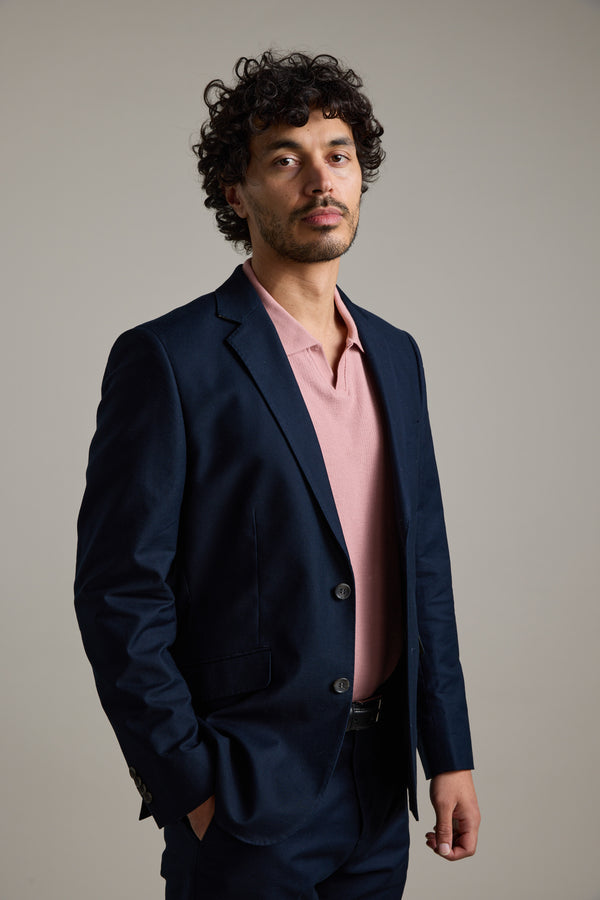 A man with curly dark hair and light facial hair wears the Barkers Moris Cotton Blazer, a navy tailored jacket, over a light pink polo. He stands confidently before a plain light gray background, looking at the camera.