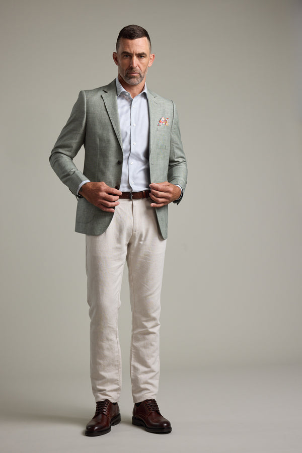 A man stands facing forward in a Barkers Marion Texture Blazer, striped dress shirt, light beige trousers, and dark brown dress shoes against a neutral grey background—a perfect summer occasion outfit.
