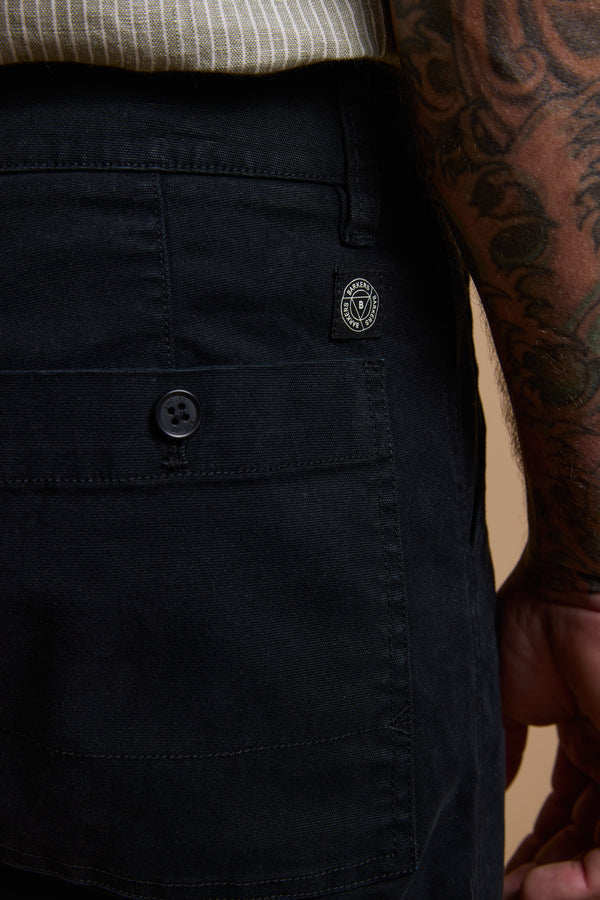 Close-up of a person wearing Barkers Tidal GD Utility Short—modern fit, black, with a buttoned pocket and small patch logo. The look is completed by a tattooed arm and striped shirt in comfortable garment-dyed cotton.