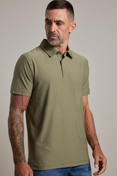 A man with short brown hair and a beard, wearing the Barkers Oakbrook Mercerised Polo in olive green and blue jeans, stands against a plain light background. He has a tattoo sleeve on his left arm and looks slightly to his left.