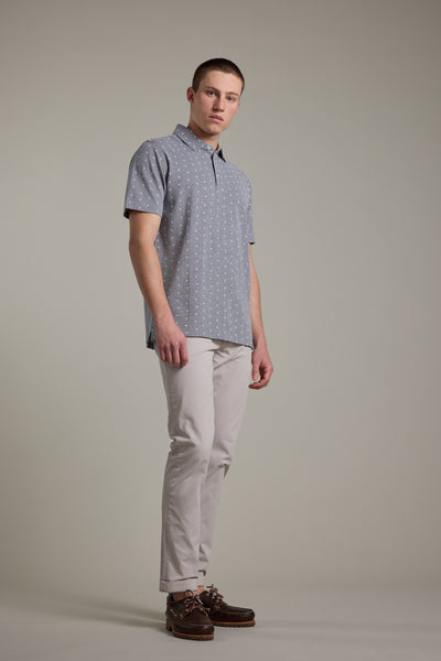 A man stands against a plain background wearing the Barkers Melrose Pique Polo, featuring a short-sleeve palm tree print in light blue, paired with light beige pants and brown boat shoes for an effortless summer look.