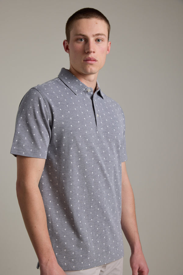 A young man with short hair models the Barkers Melrose Pique Polo in light gray with a subtle white dot pattern, set against a plain background—an ideal choice for effortless summer style.