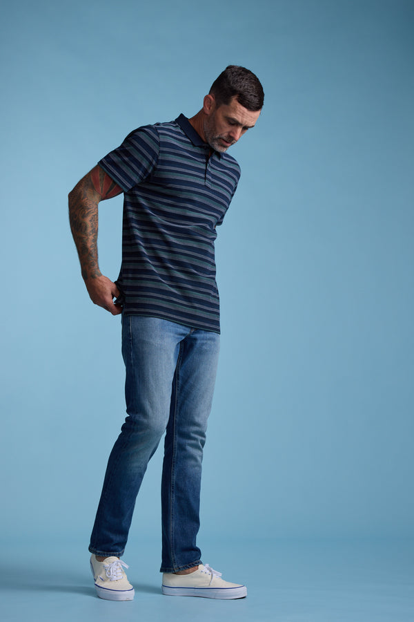 A man with short dark hair and a tattooed right arm wears the Barkers Dalton Stripe Polo, blue jeans, and white sneakers. Posing against a light blue background as he adjusts his outfit, he showcases an ideal summer look.