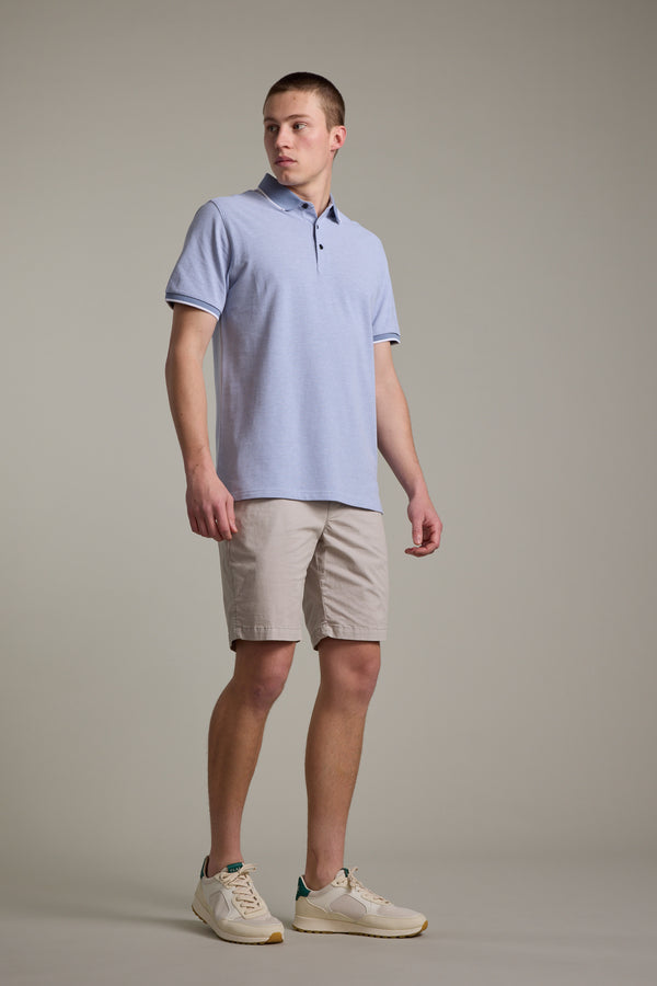 A young man stands against a plain background wearing the Barkers Birdie Pique Polo in light blue, paired with beige shorts and white sneakers. He looks slightly to the side, one hand relaxed by his side and the other at his thigh.