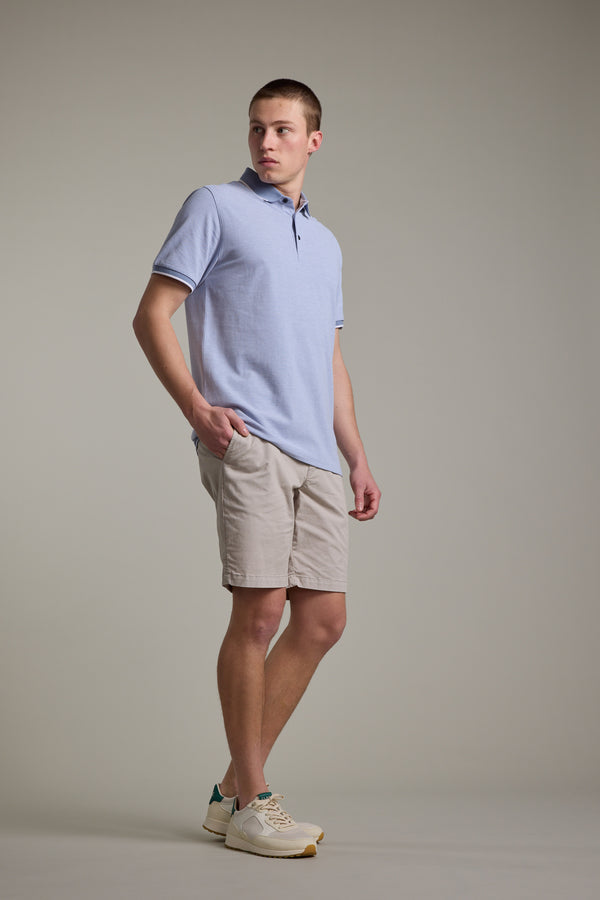 A young man stands against a plain background, wearing the Barkers Birdie Pique Polo in light blue, paired with beige shorts and cream sneakers. One hand is in his pocket as he looks slightly to the side with a neutral expression.