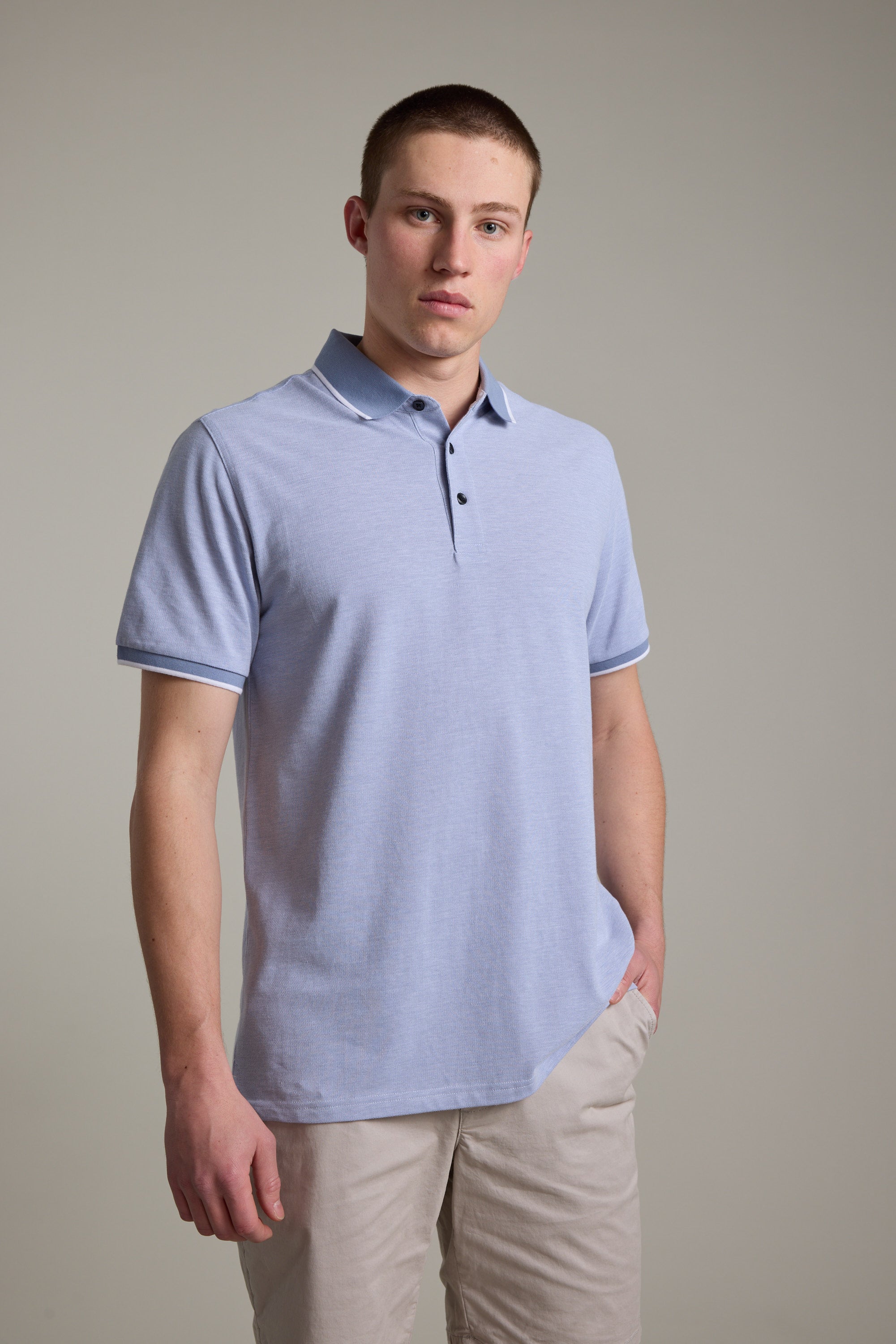 A young man with short hair wears the Barkers Birdie Pique Polo in light blue and beige shorts, standing against a plain, light-colored background with one hand in his pocket.