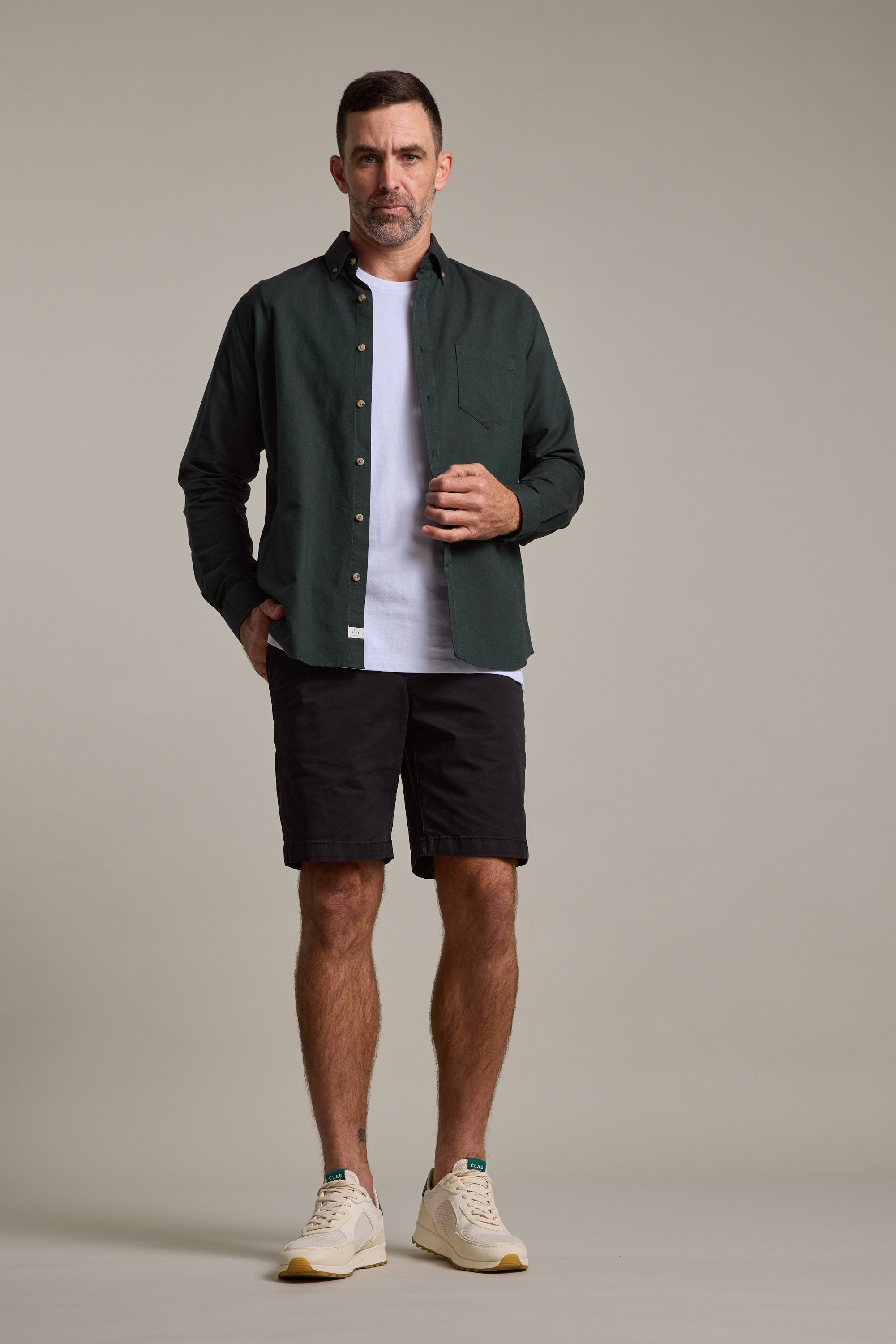 A man faces forward in a dark green button-up over a white tee, wearing Barkers Kingly Chino Shorts in black and white sneakers. The background is plain and light-colored.