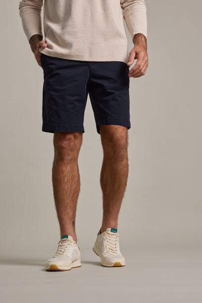 A man, shown from shoulders down against a plain background, wears a beige long-sleeve shirt and Barkers Kingly Chino Short in navy blue, paired with white sneakers featuring green accents.