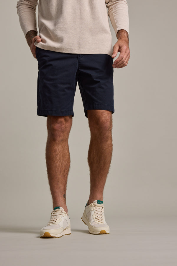 A man, shown from shoulders down against a plain background, wears a beige long-sleeve shirt and Barkers Kingly Chino Short in navy blue, paired with white sneakers featuring green accents.