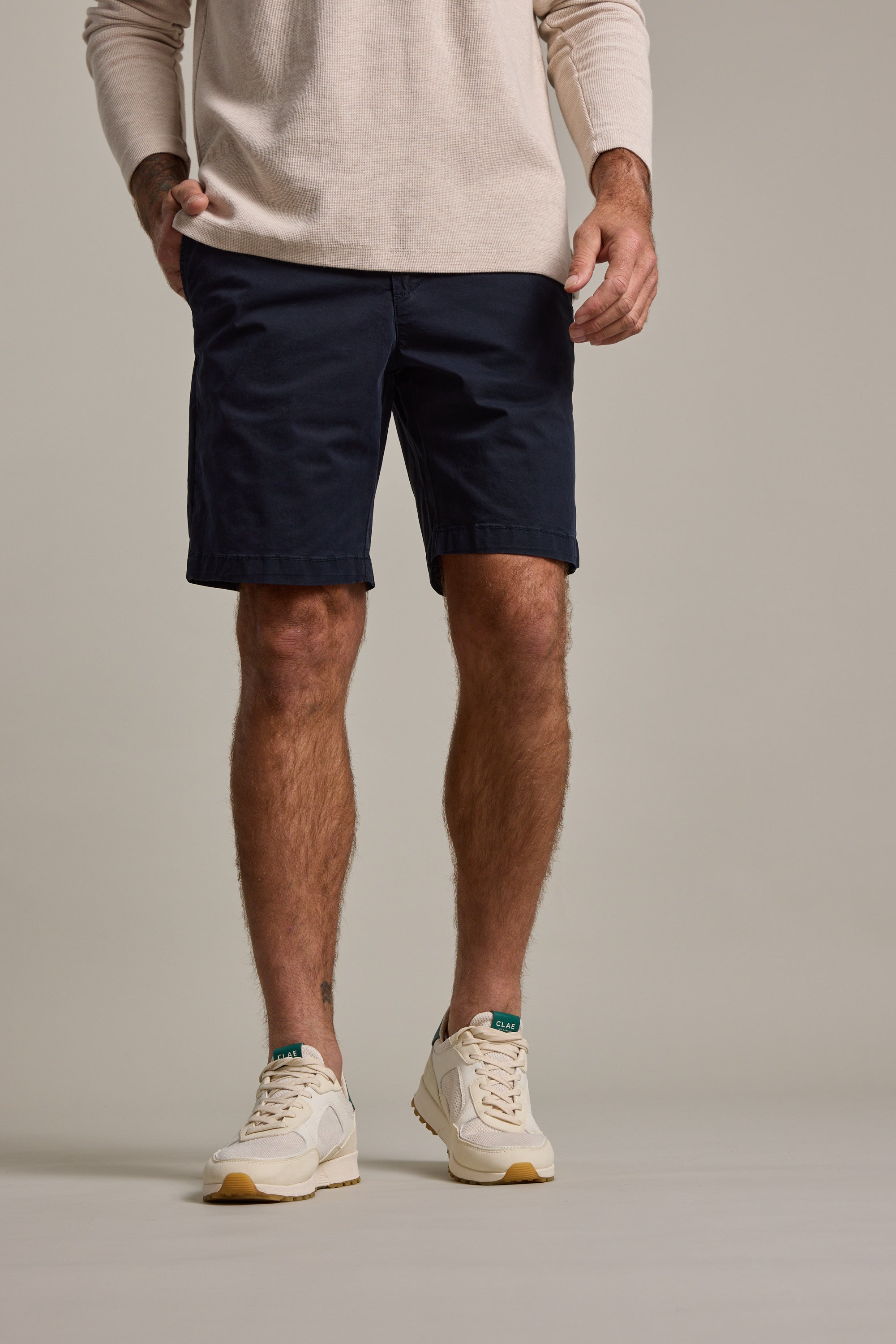 A man, shown from shoulders down against a plain background, wears a beige long-sleeve shirt and Barkers Kingly Chino Short in navy blue, paired with white sneakers featuring green accents.