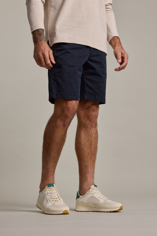 Only his lower body is visible as a man stands against a plain background wearing Barkers Kingly Chino Short—stylish men's stretch shorts in navy—paired with a beige long-sleeve shirt and white sneakers with green accents.
