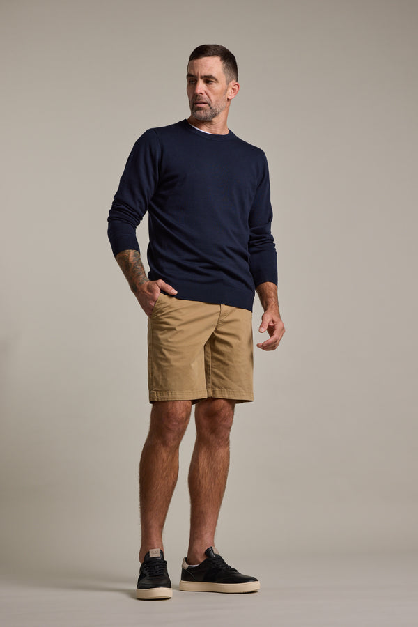 A man with short hair and a trimmed beard stands against a plain backdrop, wearing Barkers’ Kingly Chino Short—men's stretch shorts made from organic cotton—paired with a navy sweater and black sneakers, looking left with one hand in his pocket.