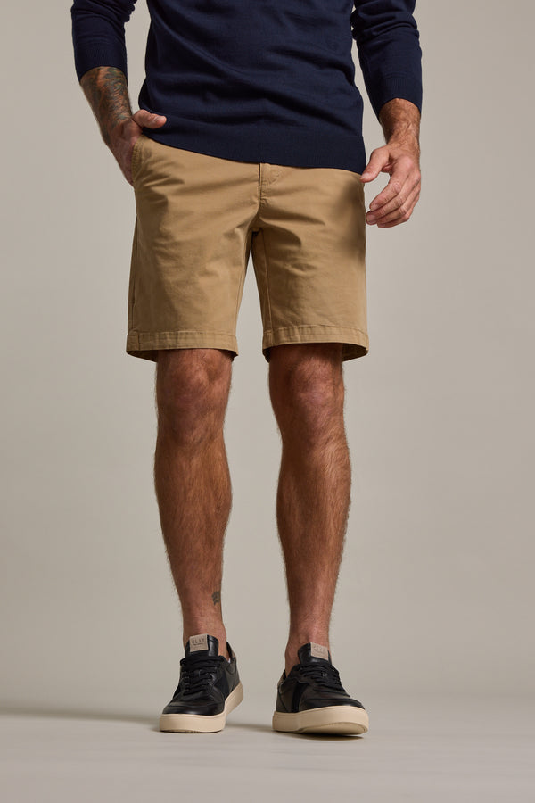 Someone stands against a beige background, hands in pockets, wearing Barkers Kingly Chino Short in tan, a navy sweater, and black sneakers with white soles. The image shows only the lower body—upper body and head are out of frame.