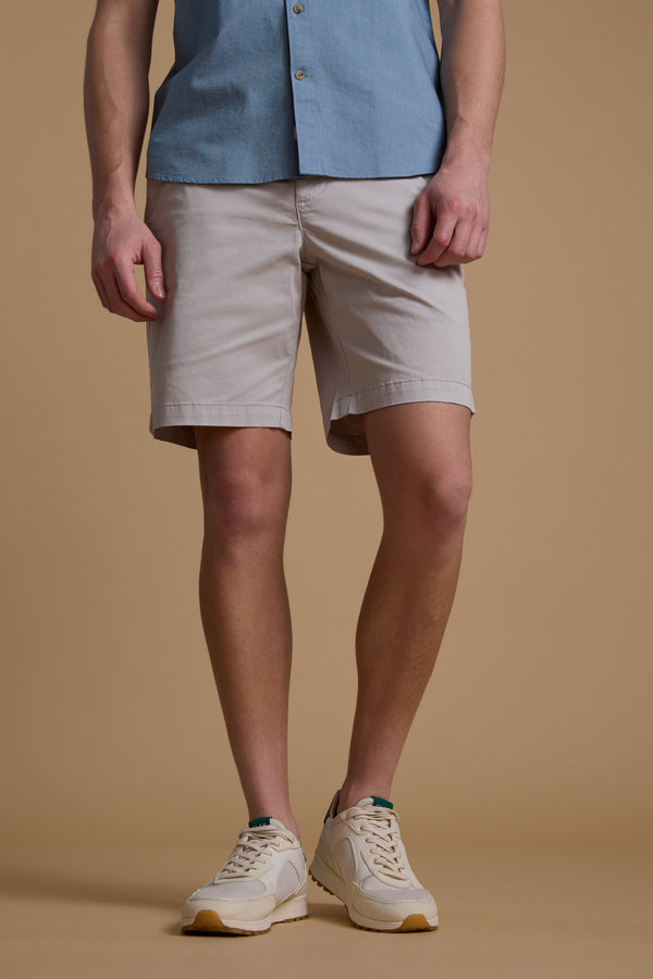 A person, only their lower half visible, stands against a tan background wearing Barkers Kingly Chino Short men's stretch shorts, beige sneakers, and a light blue short-sleeve button-up shirt.
