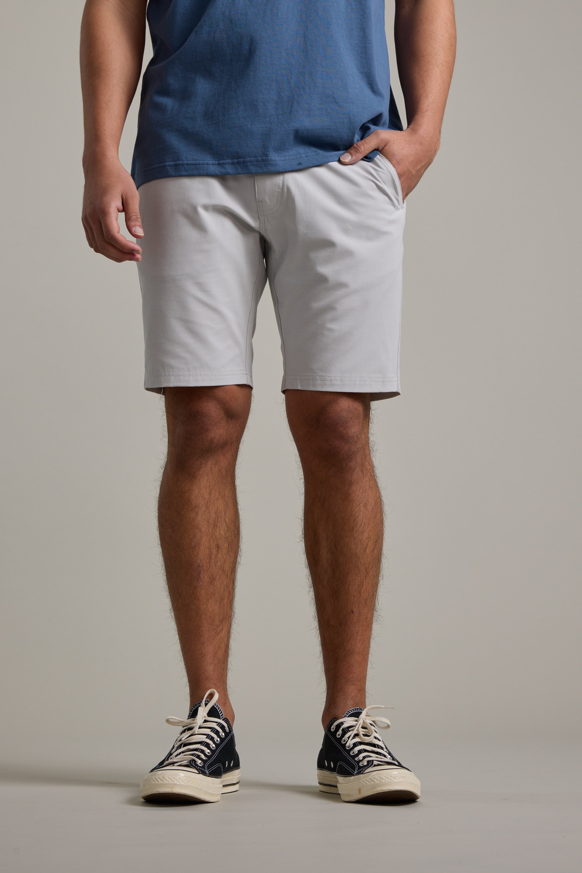 A person stands with one hand in their pocket, wearing a blue T-shirt and Barkers Hutchinson Walkshort light gray quick-dry shorts, paired with black sneakers featuring white laces. Image cropped from shoulders to feet.