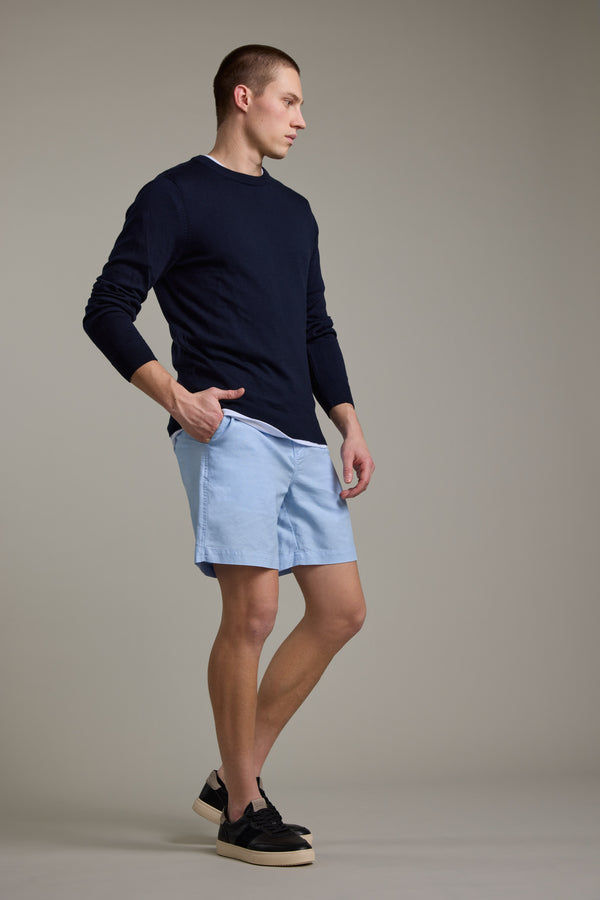 A person stands against a plain background, wearing Barkers Jasper Linen Weekend Shorts in light blue, a navy long-sleeve shirt, and black sneakers with white soles. They have short hair and are posing with one hand in their pocket.