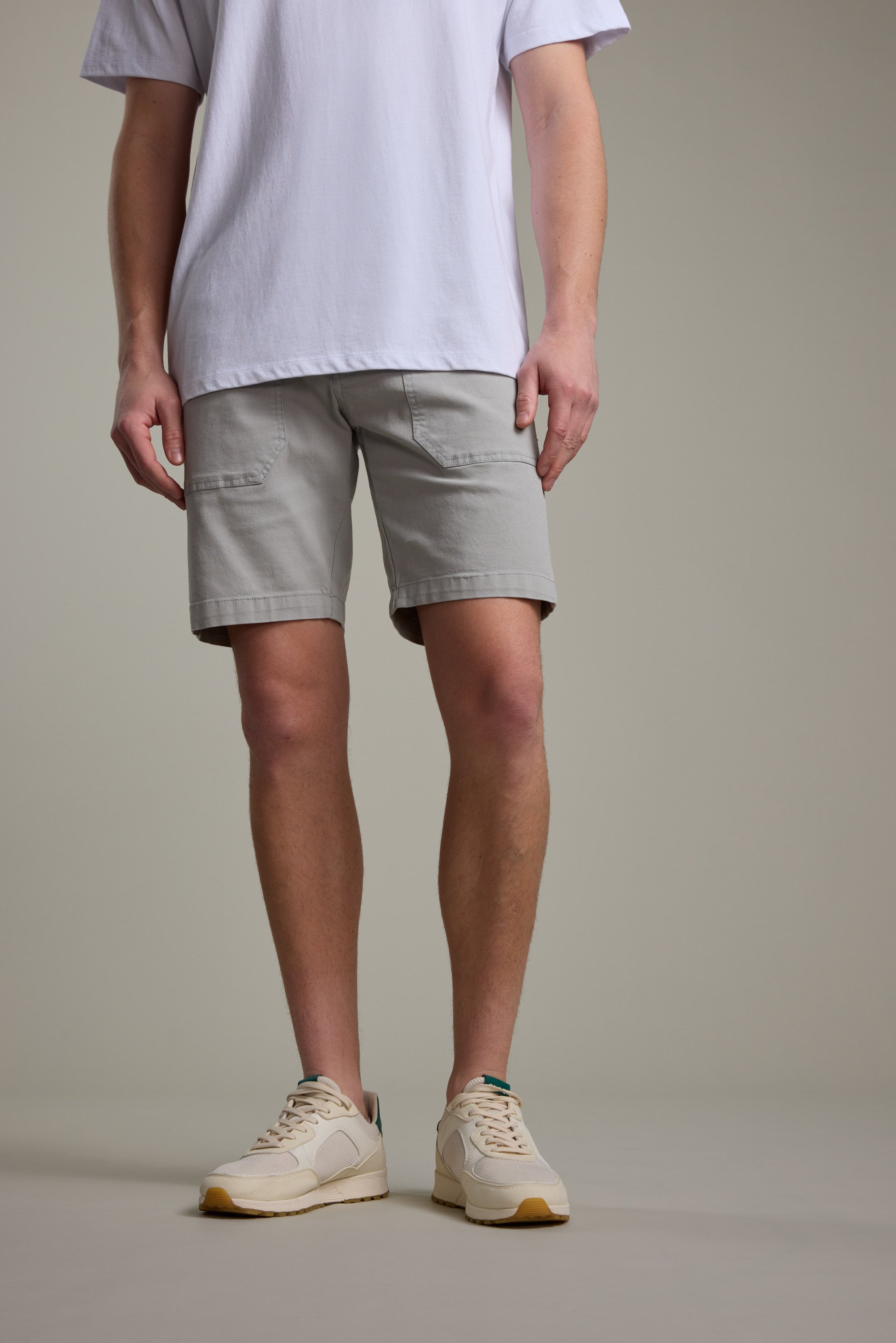 A person is shown from the shoulders down against a plain background, wearing a white t-shirt, Barkers Tidal GD Utility Shorts in light gray, and beige sneakers.