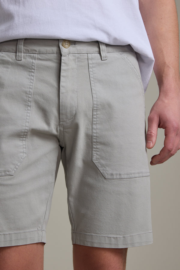 A person wearing Barkers Tidal GD Utility Shorts in light gray—featuring garment-dyed fabric, front patch pockets, and a white waistband button—with a tucked white t-shirt. Only the torso and upper legs are shown.