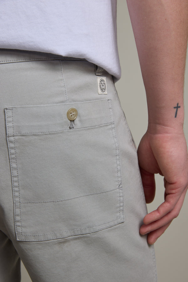 A close-up of a person’s lower back and hand, wearing Barkers Tidal GD Utility Shorts in light gray with a back pocket and beige button. A small cross tattoo is visible on their wrist, paired with a white shirt.