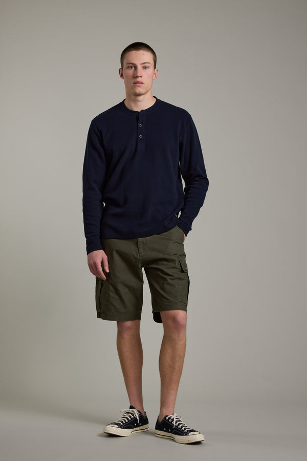 A young man stands against a plain background, wearing Barkers' Avenue Waffle Henley in dark navy, paired with olive cargo shorts and black-and-white sneakers. One hand is in his pocket as he looks straight ahead.