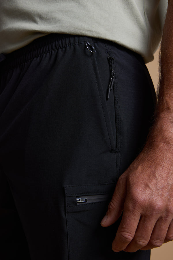 A close-up shows a person in Legasea Weekend Shorts by Legasea, featuring black four-way stretch fabric, zippered pockets, and an elastic waistband, with their right hand at their side and a light-colored shirt partially tucked in.