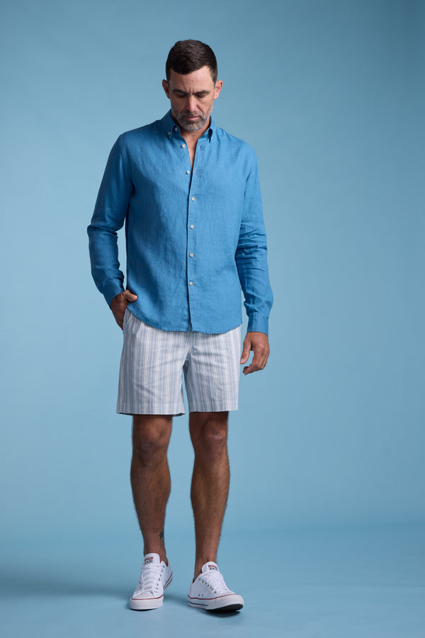A man wearing a blue button-up shirt, Barkers Manor Stripe Weekend Short with an elasticated waistband, and white sneakers stands against a blue background with one hand in his pocket, looking downward.