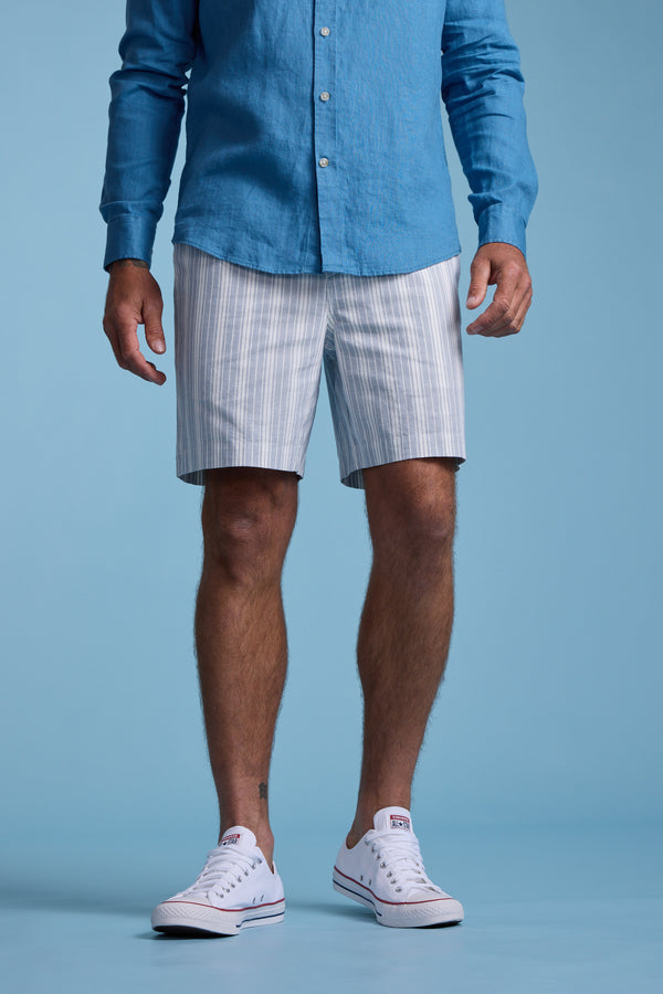 A man stands against a blue background, wearing a blue long-sleeve shirt, Barkers Manor Stripe Weekend Short with an elasticated waistband, and white sneakers. His face is not visible.