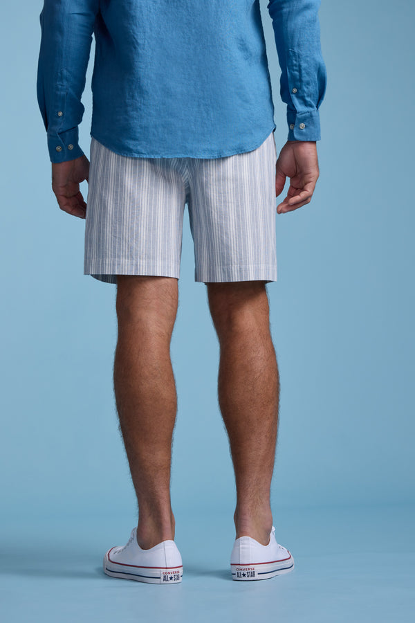 A person stands facing away, wearing a blue long-sleeve shirt, Barkers Manor Stripe Weekend Short with an elasticated waistband, and white low-top sneakers against a solid light blue background.