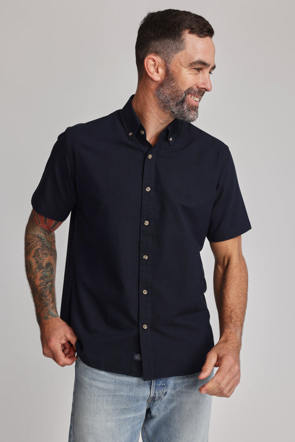 A man with short dark hair, a beard, and a tattooed arm wears the Barkers Archer Oxford Shirt in navy with a button-down collar and light blue jeans, smiling and looking to the side against a plain background.