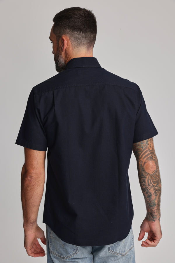 A man with short dark hair and a beard stands with his back to the camera, wearing the Barkers Archer Oxford Shirt in 100% cotton navy with a button-down collar, paired with light blue jeans. He has a tattoo sleeve on his right arm.