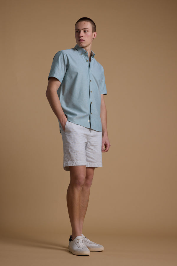 A man stands against a beige background, wearing the Barkers Archer Oxford Shirt in light blue with a button-down collar, paired with white shorts and sneakers. His hands are in his pockets as he looks to the side.