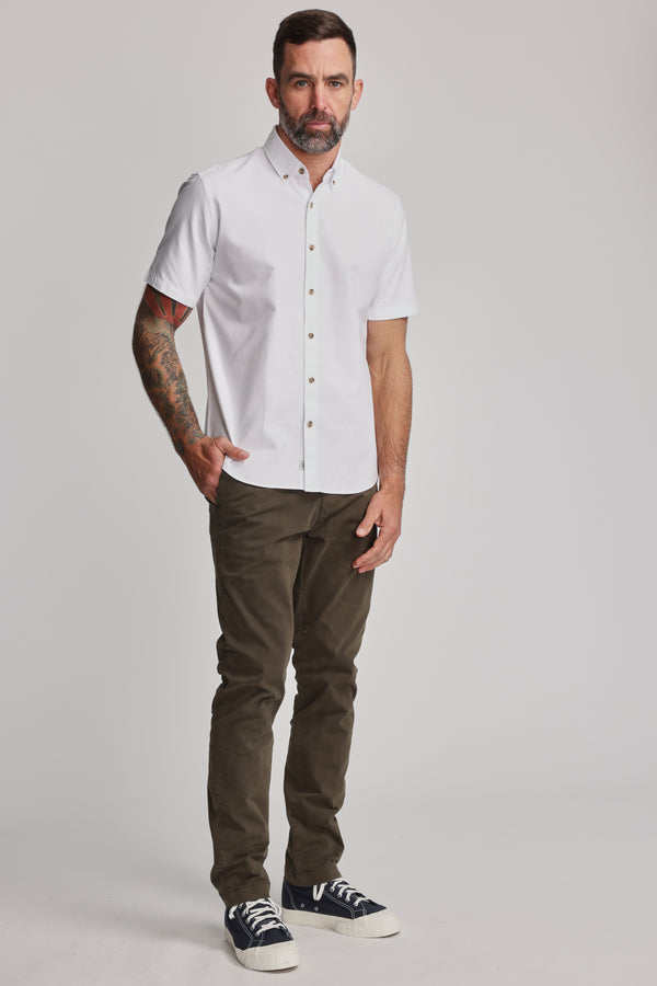 A man with short dark hair and a beard, showing tattoos on his left arm, stands against a plain background wearing the Barkers Archer Oxford Shirt in white with a button-down collar, olive green pants, and black sneakers.