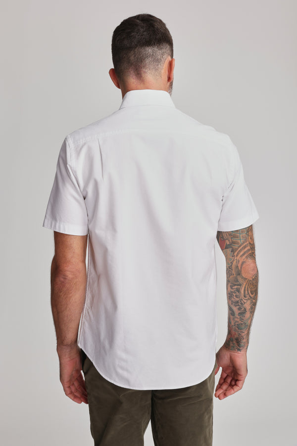 A man with short dark hair faces away, wearing Barkers' Archer Oxford Shirt (white, 100% cotton, button-down collar) and olive green pants. His right tattoo sleeve is visible against a plain light background.