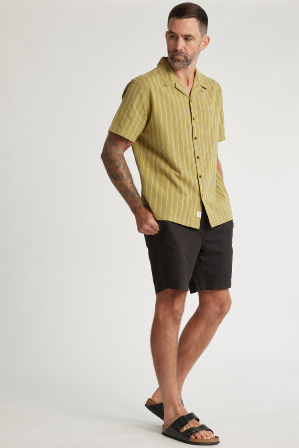 A man with short dark hair and a beard wears the Barkers Goodwin Stripe Cuban Shirt, black shorts, and black sandals. He stands with one hand in his pocket against a plain light background.