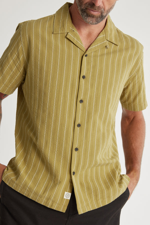 A man wears the Barkers Goodwin Stripe Cuban Shirt—a relaxed-fit, light olive green cotton shirt with white vertical stripes, short sleeves, and a camp collar—posing with one hand in his pocket against a plain background.