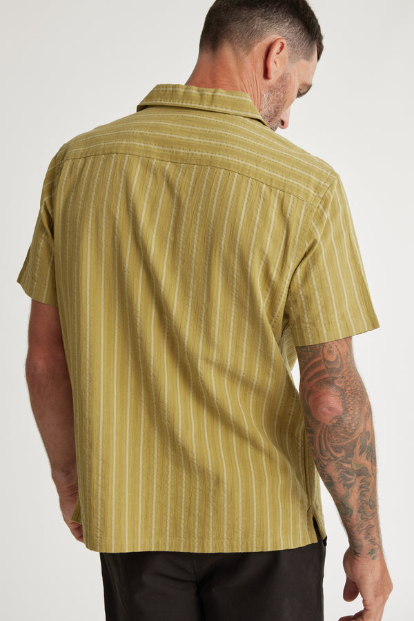 A man with short dark hair, seen from behind, wears the Barkers Goodwin Stripe Cuban Shirt—a short-sleeve cotton shirt in light olive green and white stripes. He has a tattoo sleeve on his right arm and looks slightly over his shoulder.