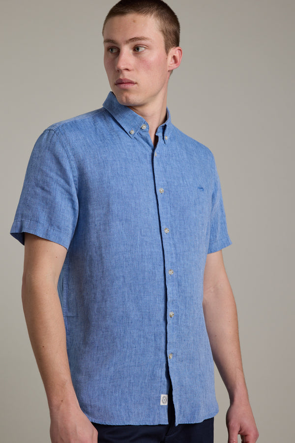 A young man with short brown hair models the Barkers Gentry Linen Shirt, a light blue, short-sleeved French linen top, paired with dark pants. He stands against a plain light background, looking to his left.