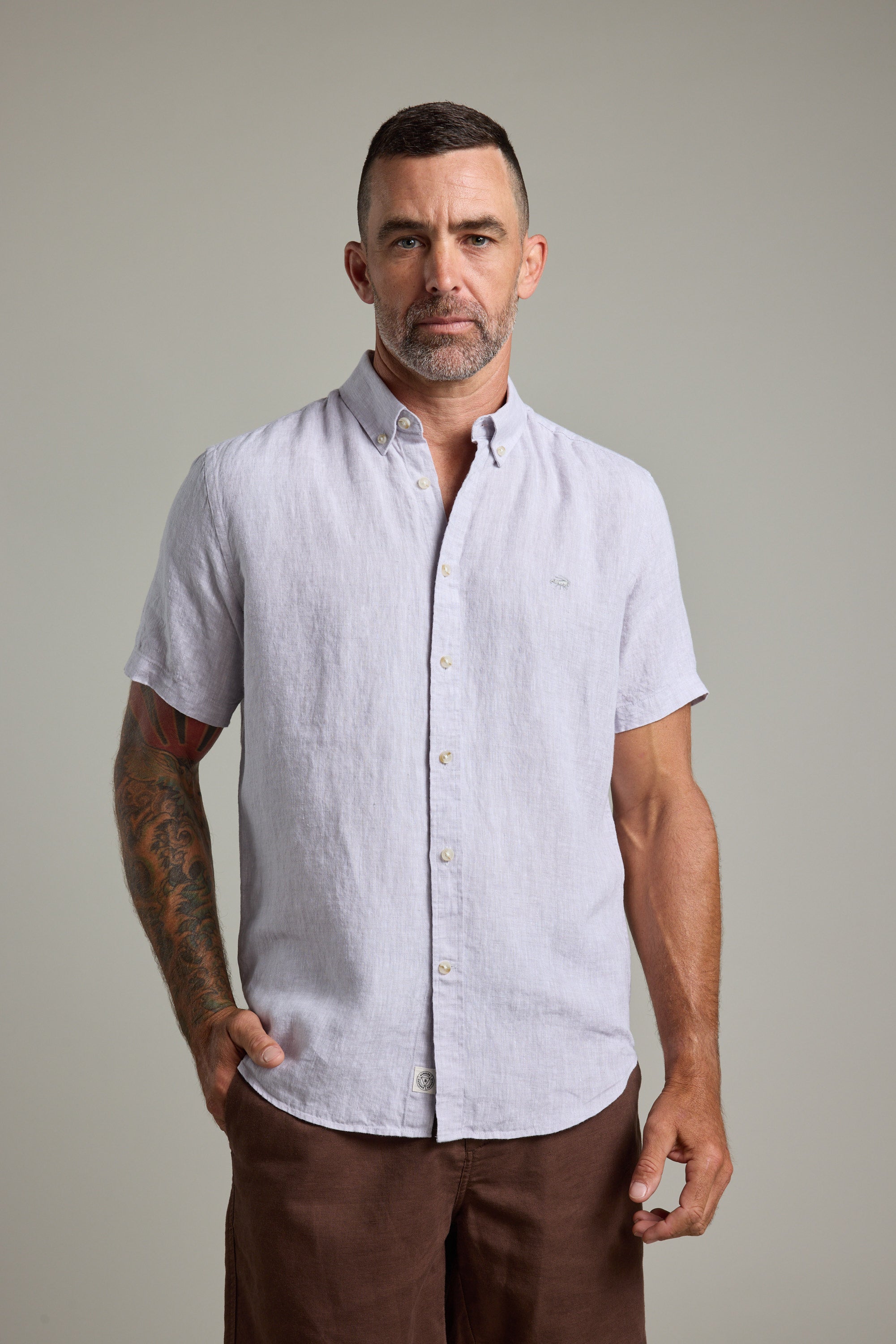 A man with short dark hair, a beard, and a tattoo sleeve on his left arm stands facing forward in Barkers' Gentry Linen Shirt (light gray, short-sleeved) and brown pants, wearing a neutral expression.