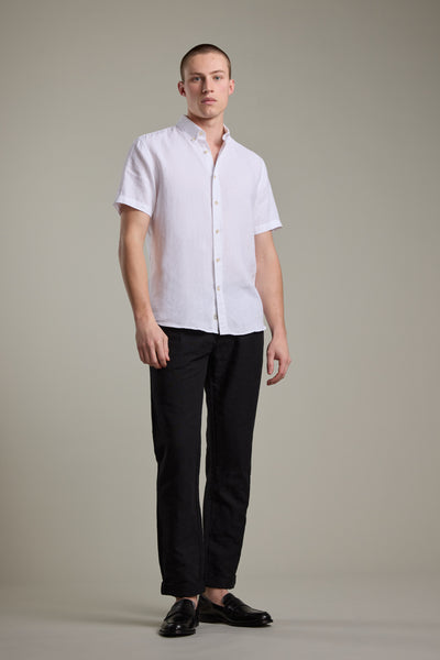 A man with short hair and a neutral expression stands against a plain background, wearing Barkers’ Gentry Linen Shirt with subtle crayfish embroidery, paired with black pants and loafers. His arms are relaxed by his sides.