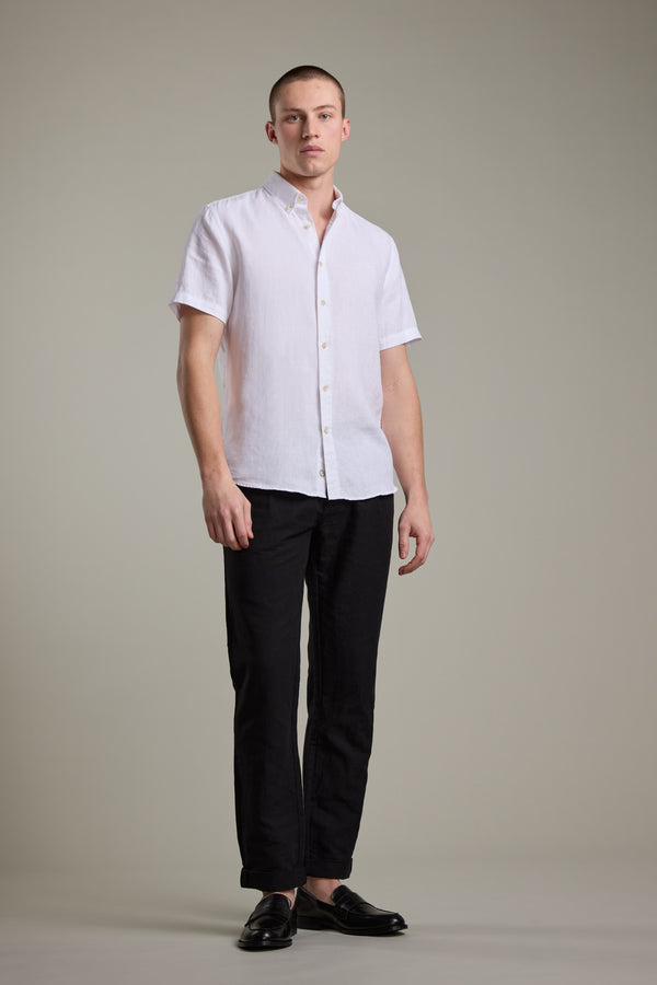 A man with short hair and a neutral expression stands against a plain background, wearing Barkers’ Gentry Linen Shirt with subtle crayfish embroidery, paired with black pants and loafers. His arms are relaxed by his sides.