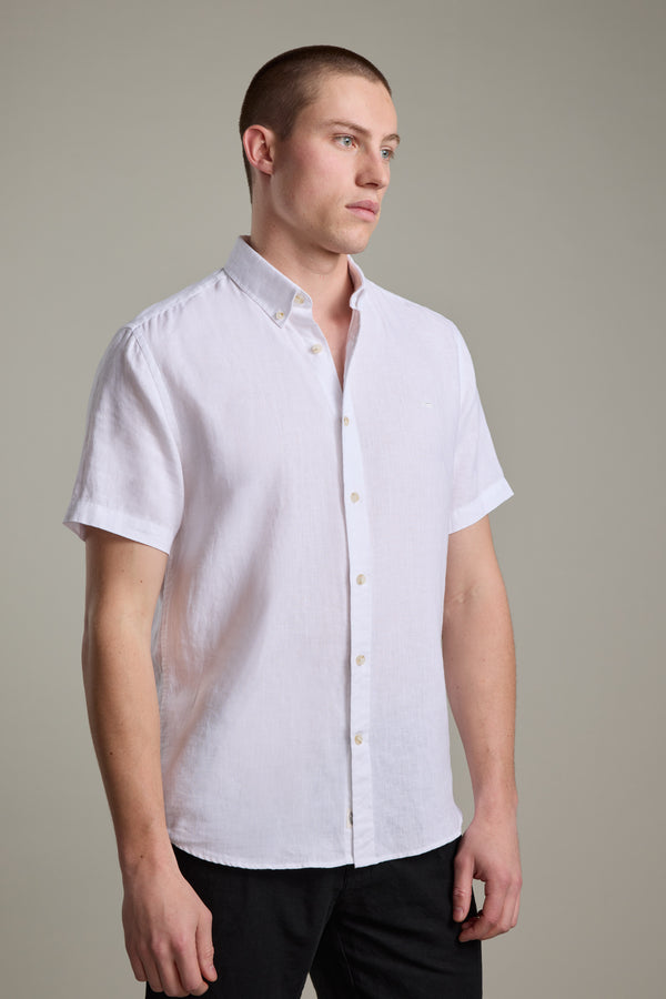 A young man with short hair wears the Barkers Gentry Linen Shirt, a short-sleeved white French linen top with subtle crayfish embroidery, paired with black pants. He stands against a plain light gray background, looking slightly to the side.