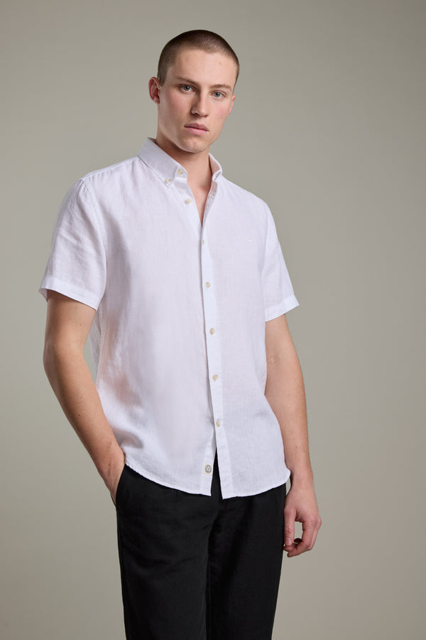 A man wears the Barkers Gentry Linen Shirt, a short-sleeved white French linen button-up, paired with black pants. He stands against a plain light gray background, hands in his pockets, looking at the camera.