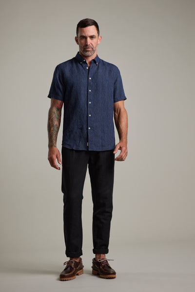A man with short dark hair and visible tattoos on his right arm stands facing forward. He is wearing the Barkers Gentry Striped Linen Shirt in navy, black pants, and brown shoes against a plain light background.