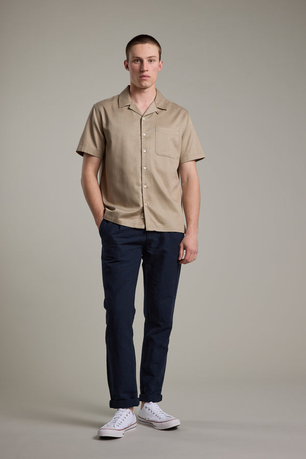 A man faces forward with a neutral expression, wearing the Barkers Augusta Cuban Shirt in tan—crafted from responsibly sourced cotton—paired with dark blue pants and white sneakers. One hand rests in his pocket against a plain background.