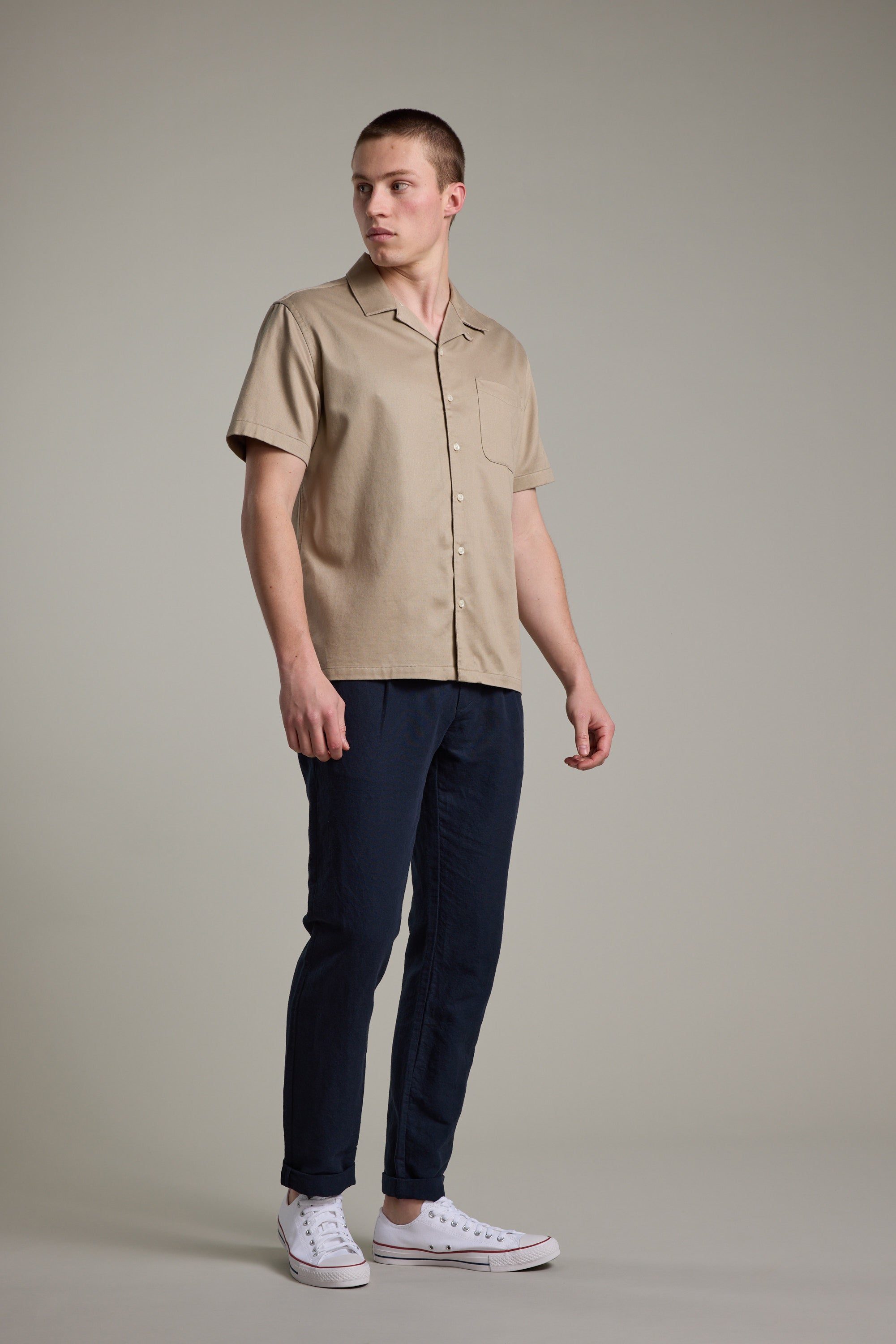 A man, looking slightly left with a neutral expression, stands against a plain background wearing the Barkers Augusta Cuban Shirt in beige—crafted from responsibly sourced cotton—with dark blue pants and white sneakers.