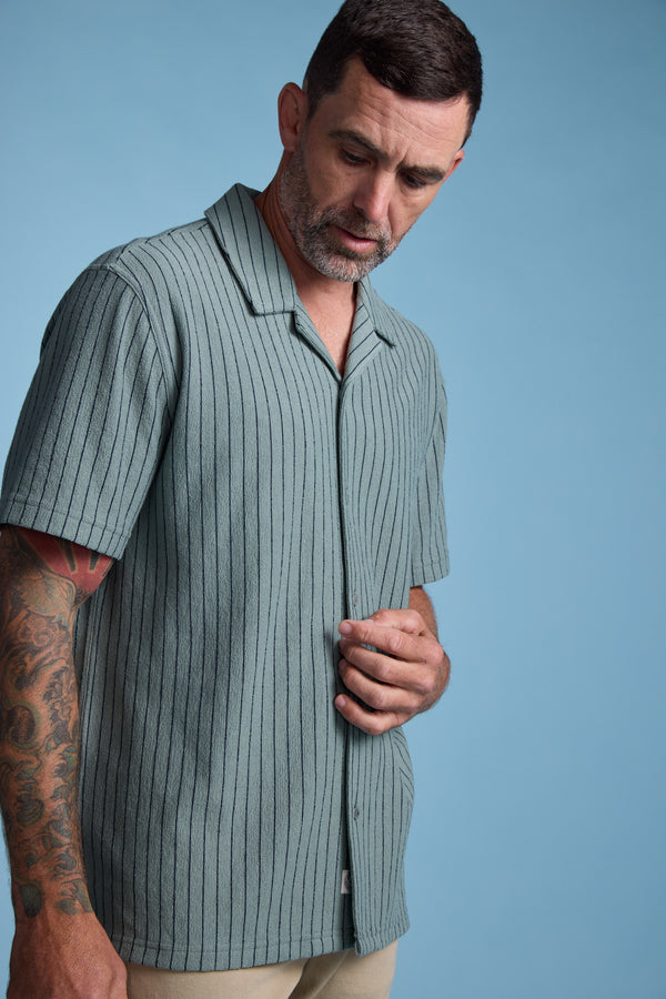 A man with short dark hair and a beard stands against a blue backdrop, wearing the Barkers Diego Stripe Cuban Shirt—a light green, vertically striped, short-sleeve design. He looks down and touches the shirt, showing tattoos on his right arm.